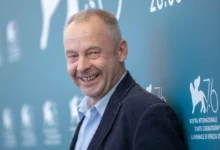 Smiling man in a suit posing against a blue wall with event signage in the background on the right side.