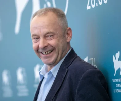 Smiling man in a suit posing against a blue wall with event signage in the background on the right side.