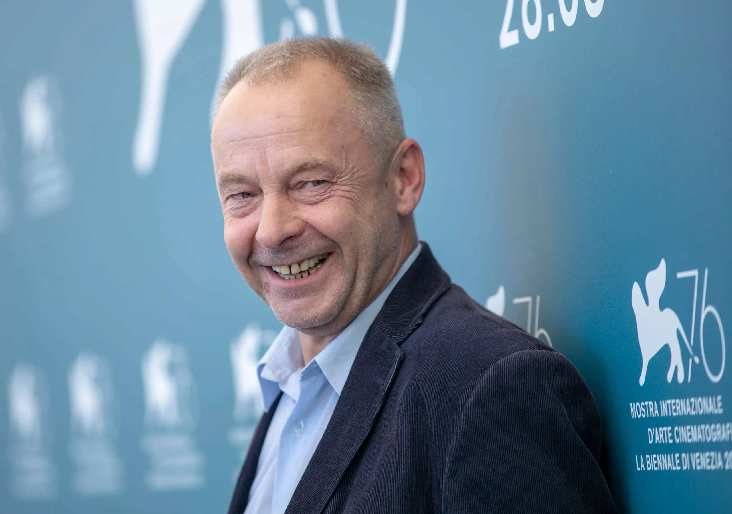 Smiling man in a suit posing against a blue wall with event signage in the background on the right side.