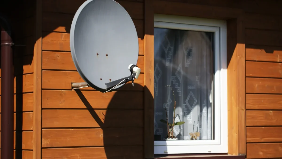Grey satellite dish mounted on the wooden exterior wall near a white-framed window of a house.