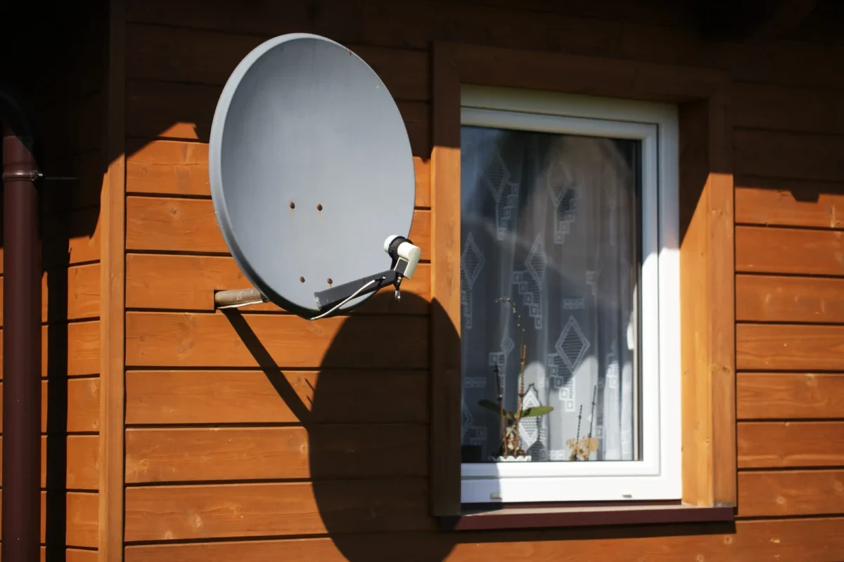 Grey satellite dish mounted on the wooden exterior wall near a white-framed window of a house.