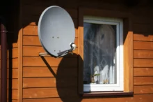 Grey satellite dish mounted on the wooden exterior wall near a white-framed window of a house.