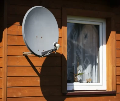 Grey satellite dish mounted on the wooden exterior wall near a white-framed window of a house.