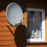 Grey satellite dish mounted on the wooden exterior wall near a white-framed window of a house.