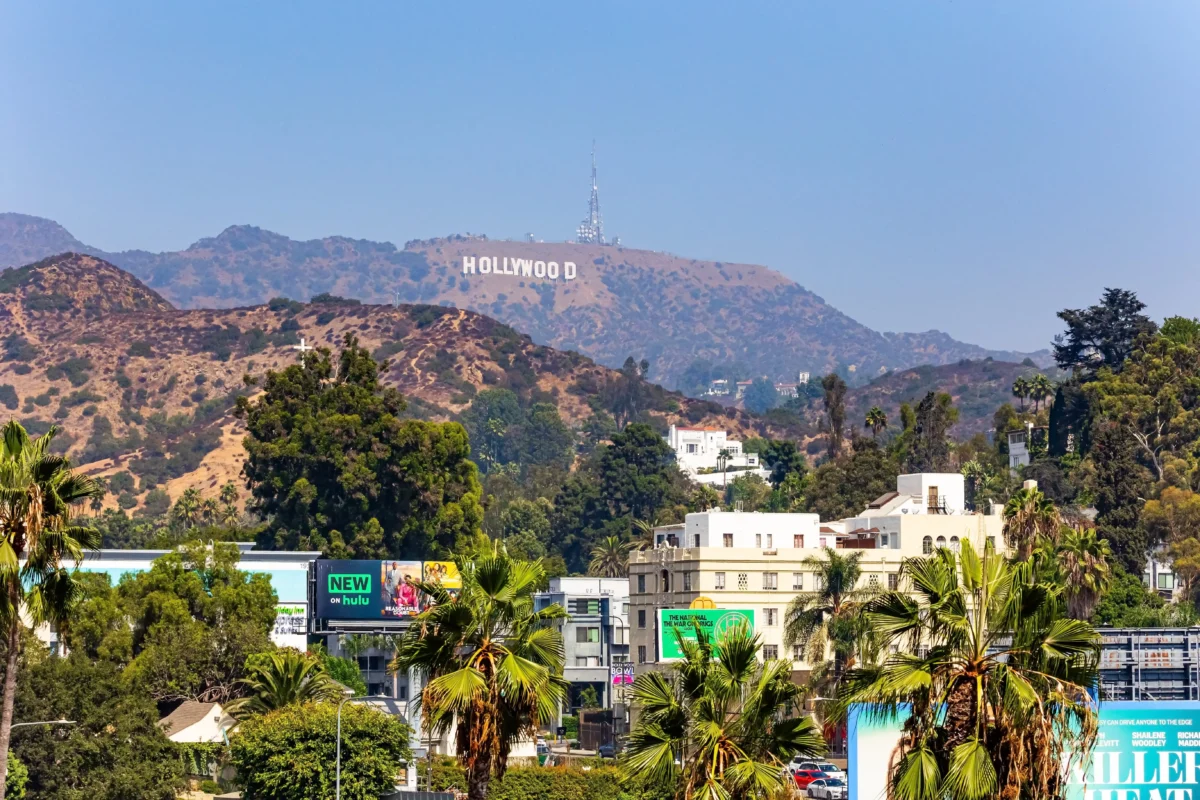 Wide hillside view with the HOLLYWOOD sign atop the hills, radio towers nearby, and an urban foreground with palm trees and billboards.