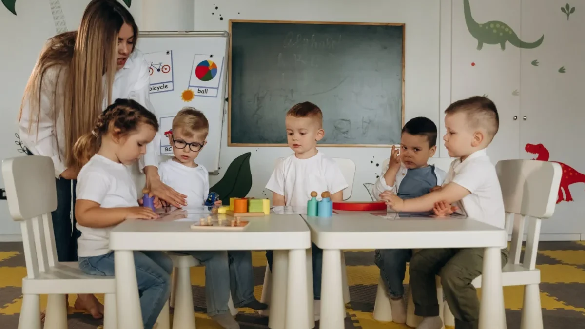 Děti ve školce u stolu a stojící učitelka