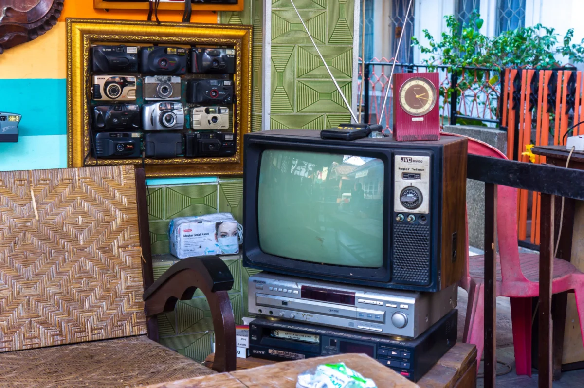 Collection of vintage cameras mounted inside a gold-framed display, with a retro TV and clock on a green patterned wall outdoors.