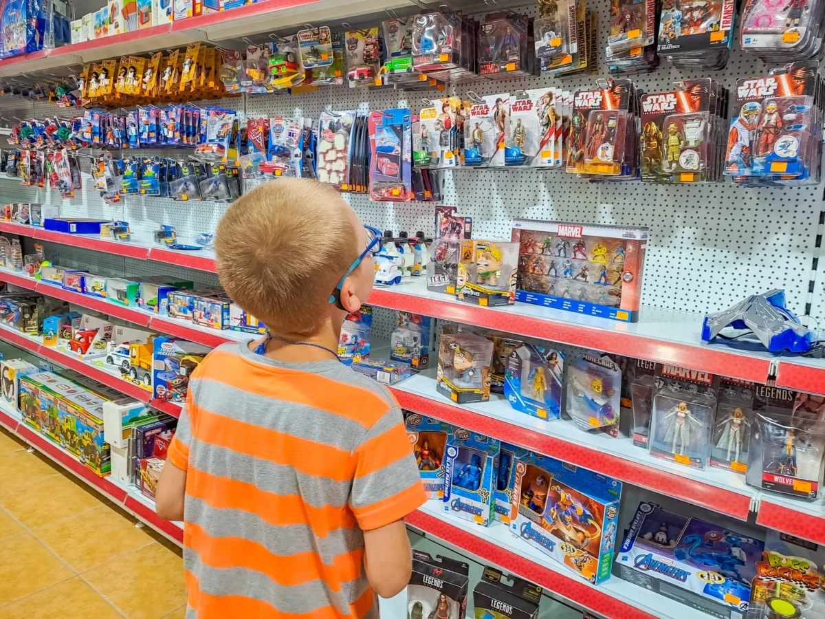 shutterstock_2661949645 1 | TVGURU.cz Young boy in an orange and gray striped shirt browsing shelves of action figures in a toy store.