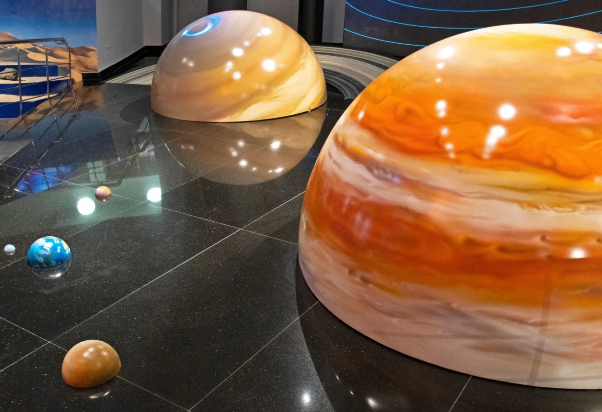 Vnitřní expozice s nadrozměrnými mramorovanými planetami na lesklé černé podlaze; modrá planeta vlevo, velká oranžová planeta vpravo.