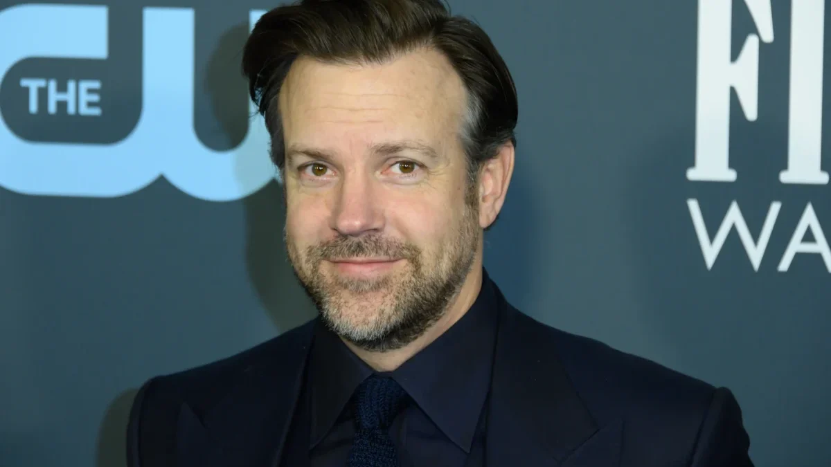 Portrait of a man in a dark suit and tie, smiling, with a blue backdrop and white lettering behind him.