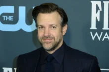 Portrait of a man in a dark suit and tie, smiling, with a blue backdrop and white lettering behind him.