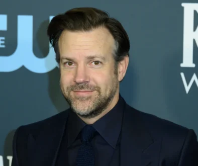 Portrait of a man in a dark suit and tie, smiling, with a blue backdrop and white lettering behind him.