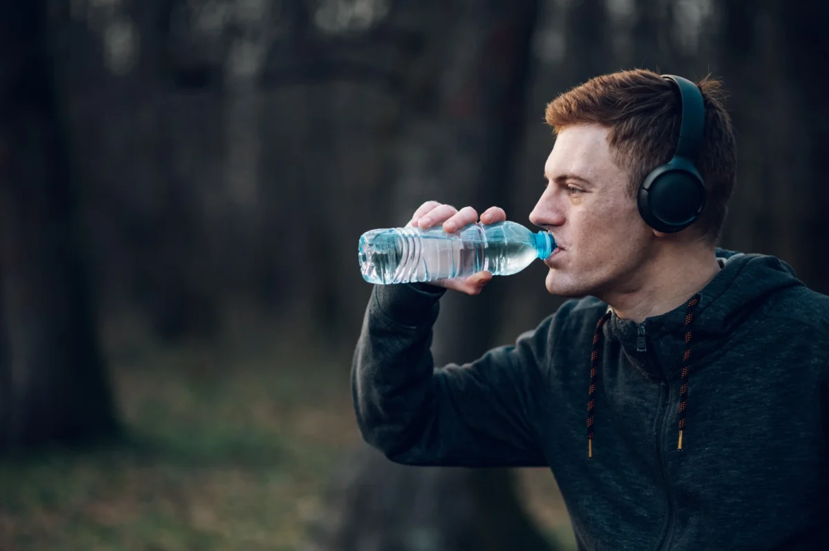 Man wearing over-ear headphones drinks from a plastic water bottle outdoors, forest in the background? (informational)