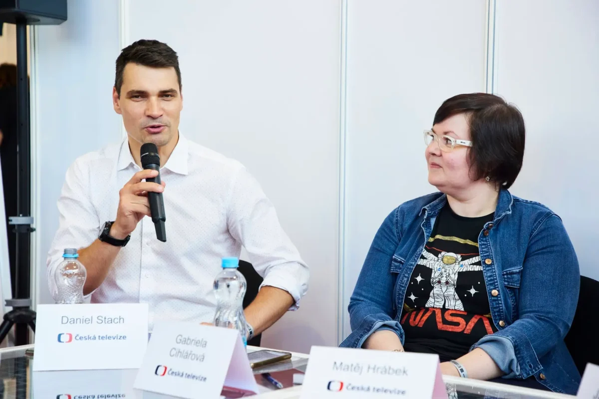 Moderátor Daniel Stach mluví do mikrofonu a vedle něj sedí žena.