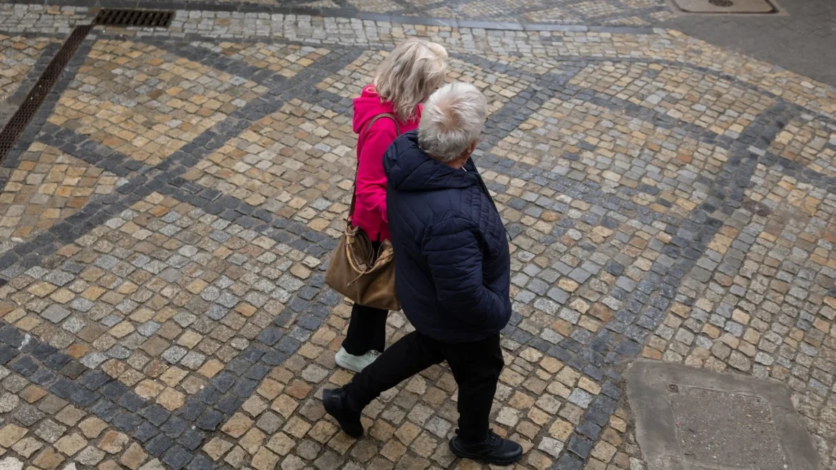 Two people walk together on a decorative beige and gray cobblestone street, seen from above; one wears a pink jacket and carries a brown bag, the other in a dark jacket.