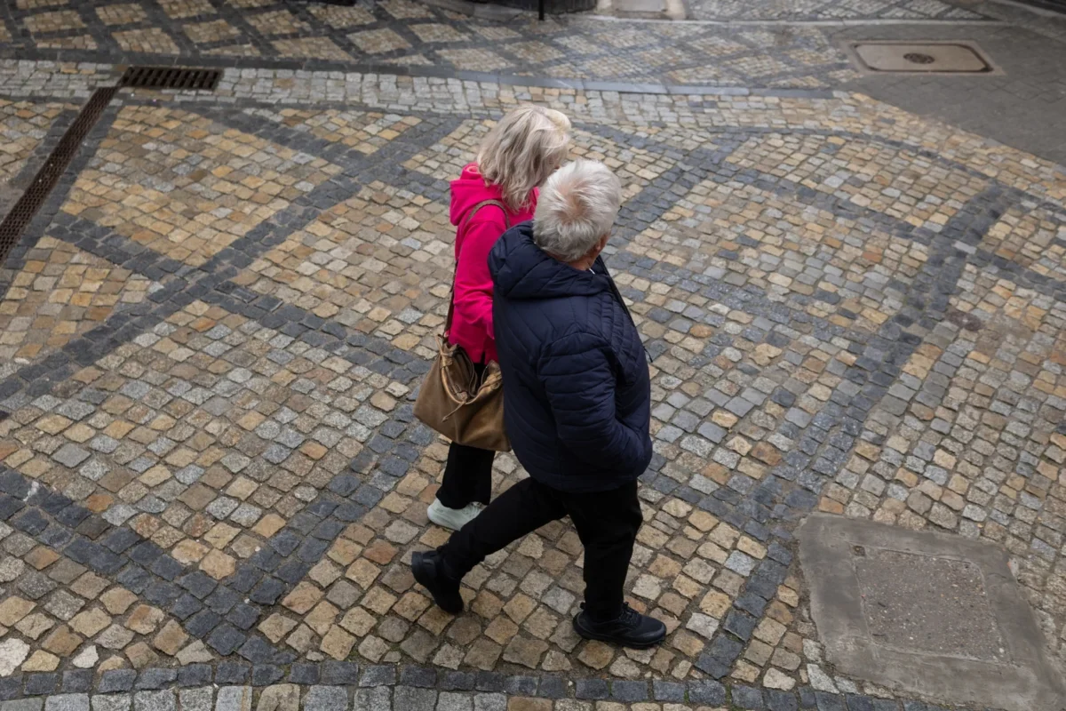 Two people walk together on a decorative beige and gray cobblestone street, seen from above; one wears a pink jacket and carries a brown bag, the other in a dark jacket.