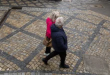 Two people walk together on a decorative beige and gray cobblestone street, seen from above; one wears a pink jacket and carries a brown bag, the other in a dark jacket.