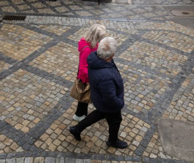 Two people walk together on a decorative beige and gray cobblestone street, seen from above; one wears a pink jacket and carries a brown bag, the other in a dark jacket.