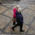 Two people walk together on a decorative beige and gray cobblestone street, seen from above; one wears a pink jacket and carries a brown bag, the other in a dark jacket.