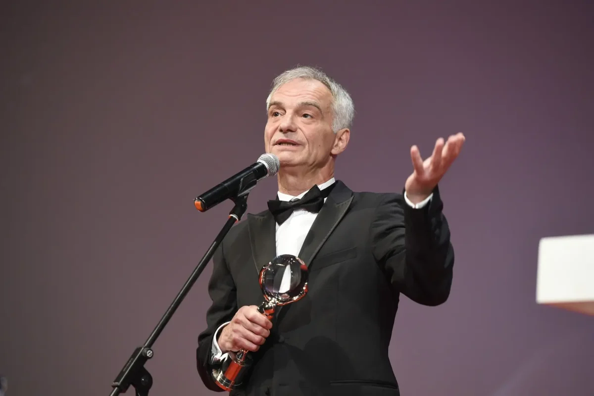 Herec Ivan Trojan v černém smokingu a motýlku mluví do mikrofonu, zatímco na pódiu drží trofej. Při mluvení natahuje jednu ruku.