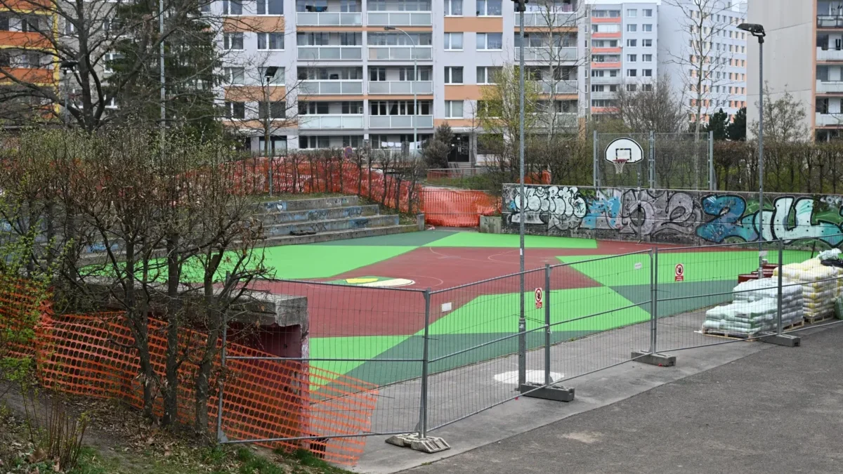 Basketball court under construction in a city area, with bright green and red playing surface, graffiti wall, orange safety fencing, and apartment buildings in the background.