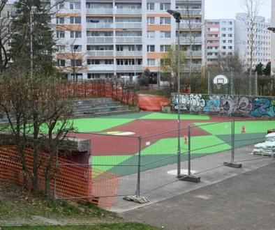 Basketball court under construction in a city area, with bright green and red playing surface, graffiti wall, orange safety fencing, and apartment buildings in the background.