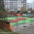 Basketball court under construction in a city area, with bright green and red playing surface, graffiti wall, orange safety fencing, and apartment buildings in the background.
