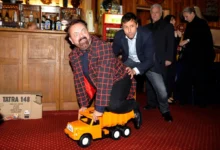 Man in a red checkered coat sits on an orange toy truck as another man nudges him from behind in a bar setting.