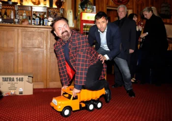 Man in a red checkered coat sits on an orange toy truck as another man nudges him from behind in a bar setting.