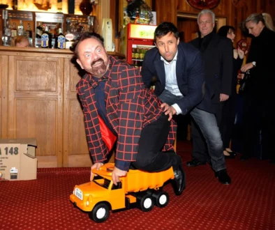 Man in a red checkered coat sits on an orange toy truck as another man nudges him from behind in a bar setting.