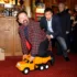 Man in a red checkered coat sits on an orange toy truck as another man nudges him from behind in a bar setting.