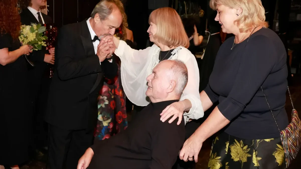 Formal ceremony: a man in a black tuxedo kisses the hands of a seated man in a wheelchair, as two women in dark attire stand nearby. Floral bouquets are visible in the background.
