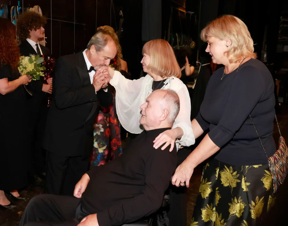 Formal ceremony: a man in a black tuxedo kisses the hands of a seated man in a wheelchair, as two women in dark attire stand nearby. Floral bouquets are visible in the background.