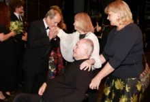Formal ceremony: a man in a black tuxedo kisses the hands of a seated man in a wheelchair, as two women in dark attire stand nearby. Floral bouquets are visible in the background.