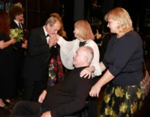 Formal ceremony: a man in a black tuxedo kisses the hands of a seated man in a wheelchair, as two women in dark attire stand nearby. Floral bouquets are visible in the background.
