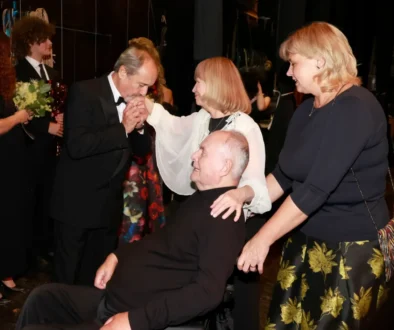 Formal ceremony: a man in a black tuxedo kisses the hands of a seated man in a wheelchair, as two women in dark attire stand nearby. Floral bouquets are visible in the background.