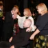 Formal ceremony: a man in a black tuxedo kisses the hands of a seated man in a wheelchair, as two women in dark attire stand nearby. Floral bouquets are visible in the background.