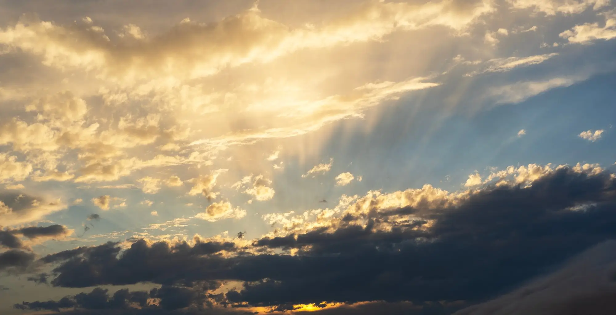 Sunset sky with golden clouds and sunbeams piercing through the clouds toward the horizon.