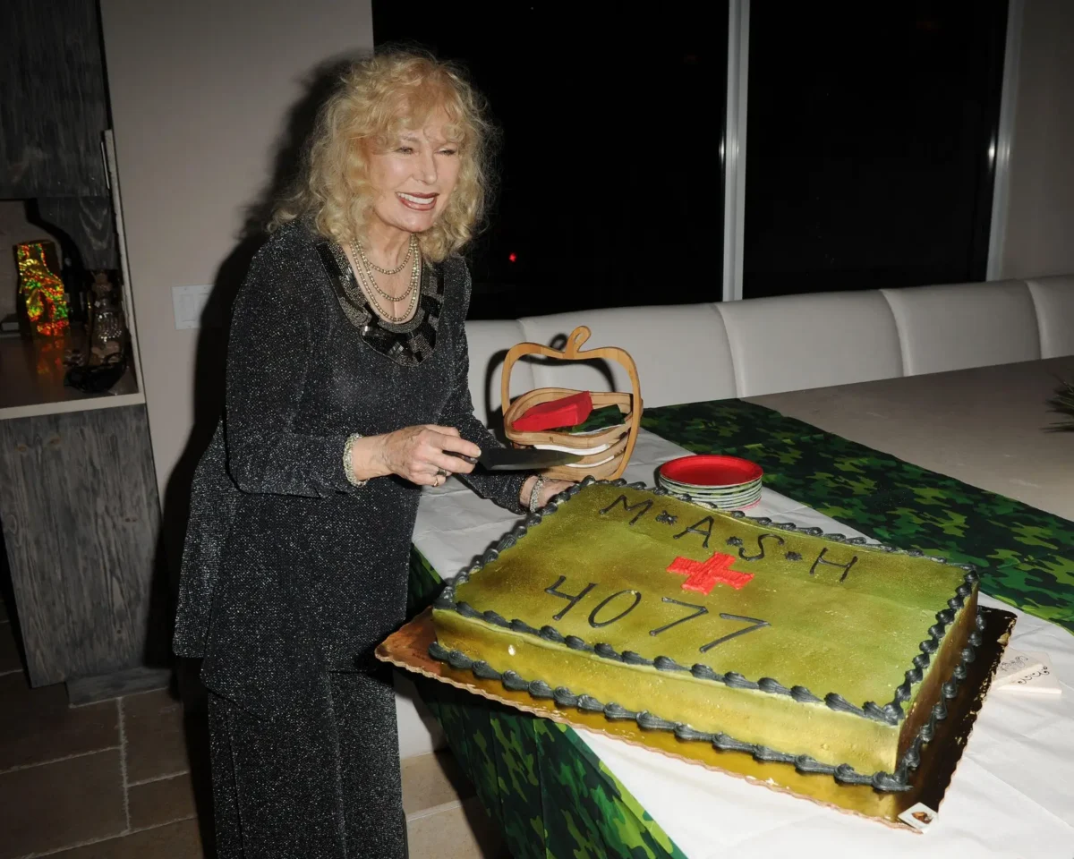 Older woman with curly blond hair in a sparkly dark dress cutting a large two‑tier gold birthday cake on a table.
