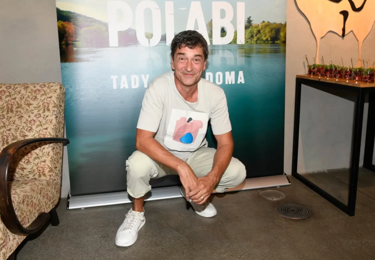 Man smiling and squatting indoors in front of a lake-and-forest backdrop with large white letters, a chair on the left and a table with drinks on the right.