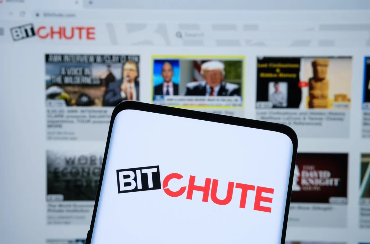 Blurred computer screen showing video thumbnails, including a prominent BITCHUTE logo in red and black, with a curved white device in the foreground.