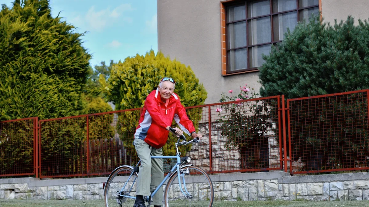 Older man in a red jacket rides a bicycle along a grassy path beside a red metal fence and a beige building with large windows.