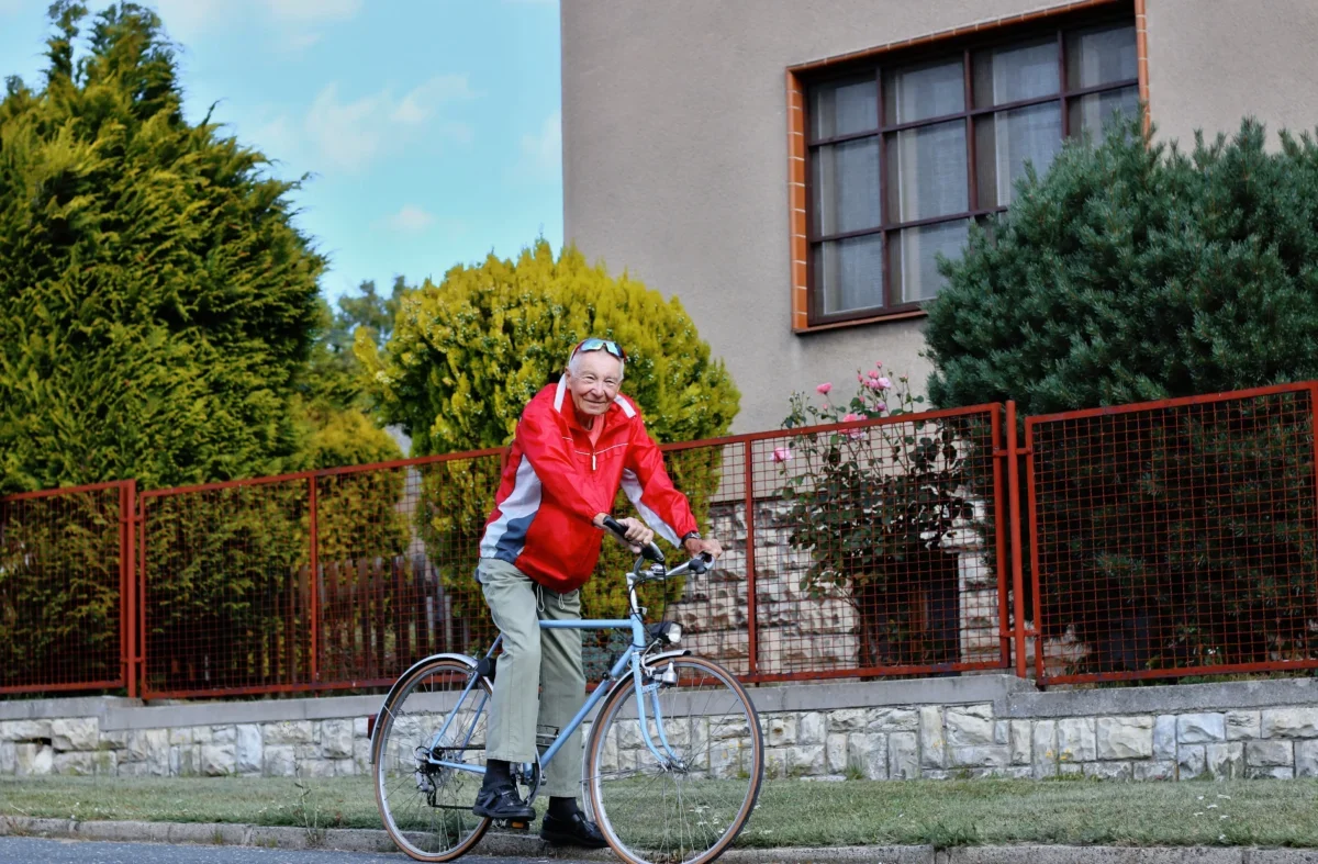 Older man in a red jacket rides a bicycle along a grassy path beside a red metal fence and a beige building with large windows.