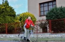 Older man in a red jacket rides a bicycle along a grassy path beside a red metal fence and a beige building with large windows.