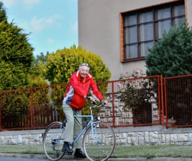 Older man in a red jacket rides a bicycle along a grassy path beside a red metal fence and a beige building with large windows.