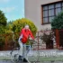 Older man in a red jacket rides a bicycle along a grassy path beside a red metal fence and a beige building with large windows.