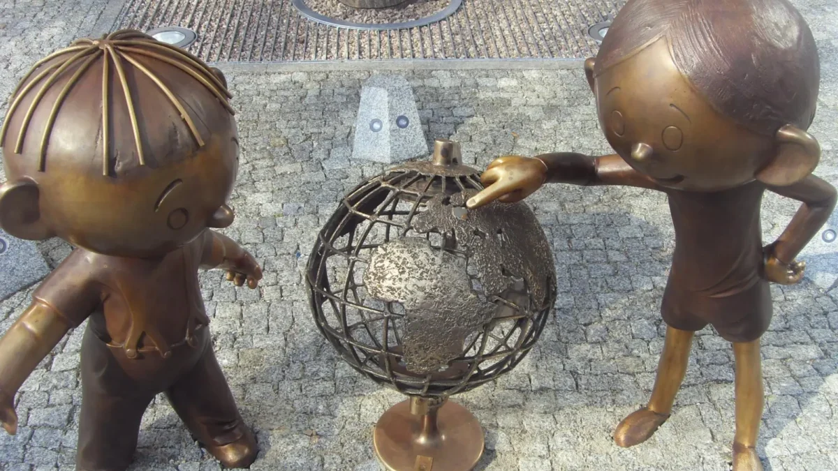 Two bronze child statues lean toward a rusted metal sphere sculpture in a plaza with a tree and cobblestone ground.