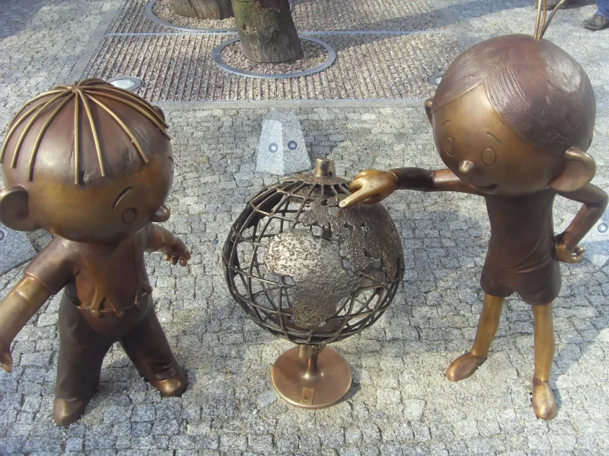 Two bronze child statues lean toward a rusted metal sphere sculpture in a plaza with a tree and cobblestone ground.