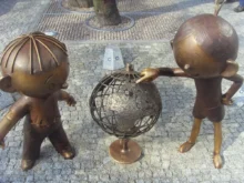 Two bronze child statues lean toward a rusted metal sphere sculpture in a plaza with a tree and cobblestone ground.