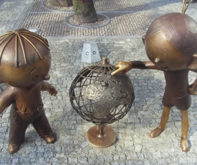 Two bronze child statues lean toward a rusted metal sphere sculpture in a plaza with a tree and cobblestone ground.